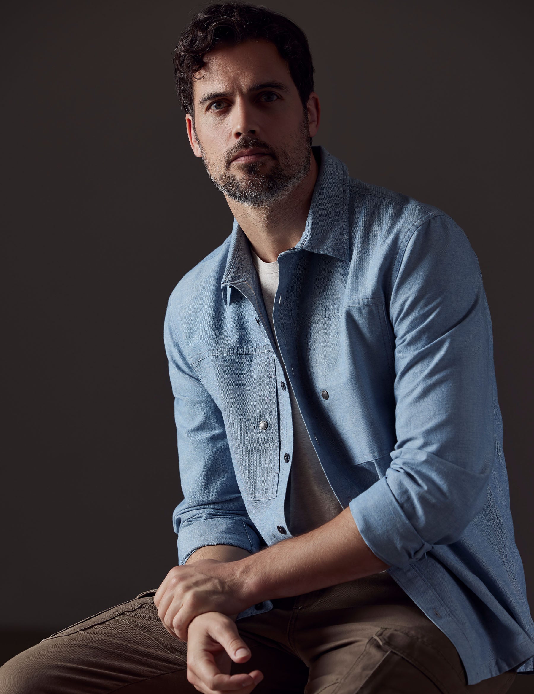 Man sitting with arms crossed wearing unbuttoned Emerson Button Down in Light Blue from AETHER Apparel.