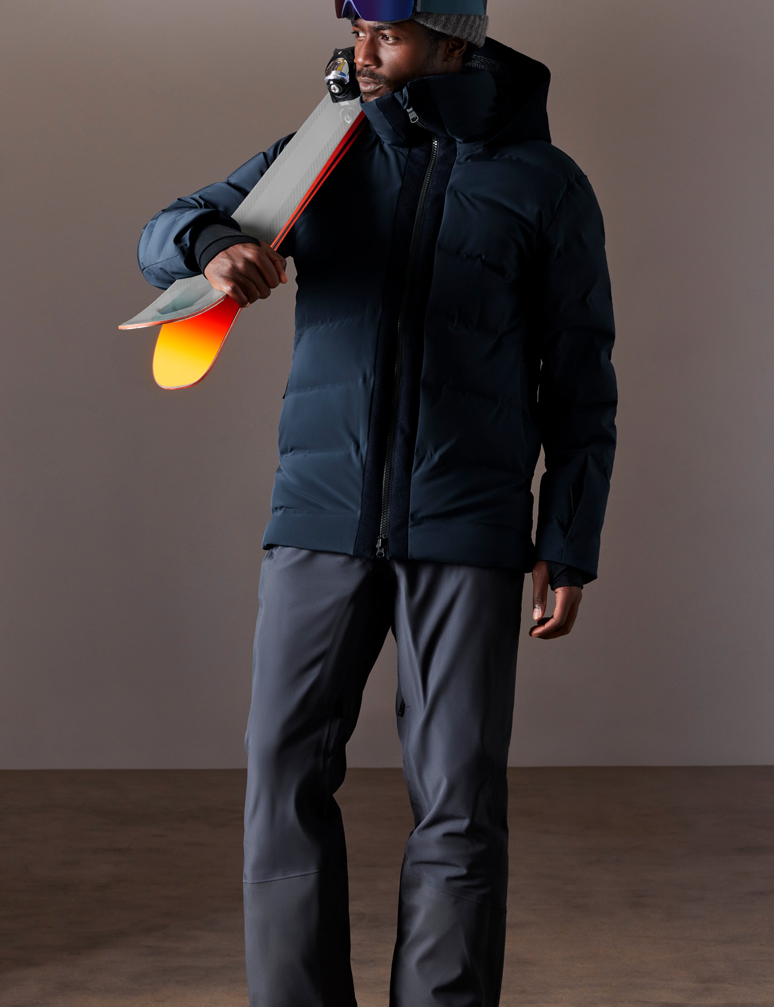 Front full-body view of man holding skis over shoulder wearing Nordic Jacket in Total Eclipse dark blue from AETHER Apparel.