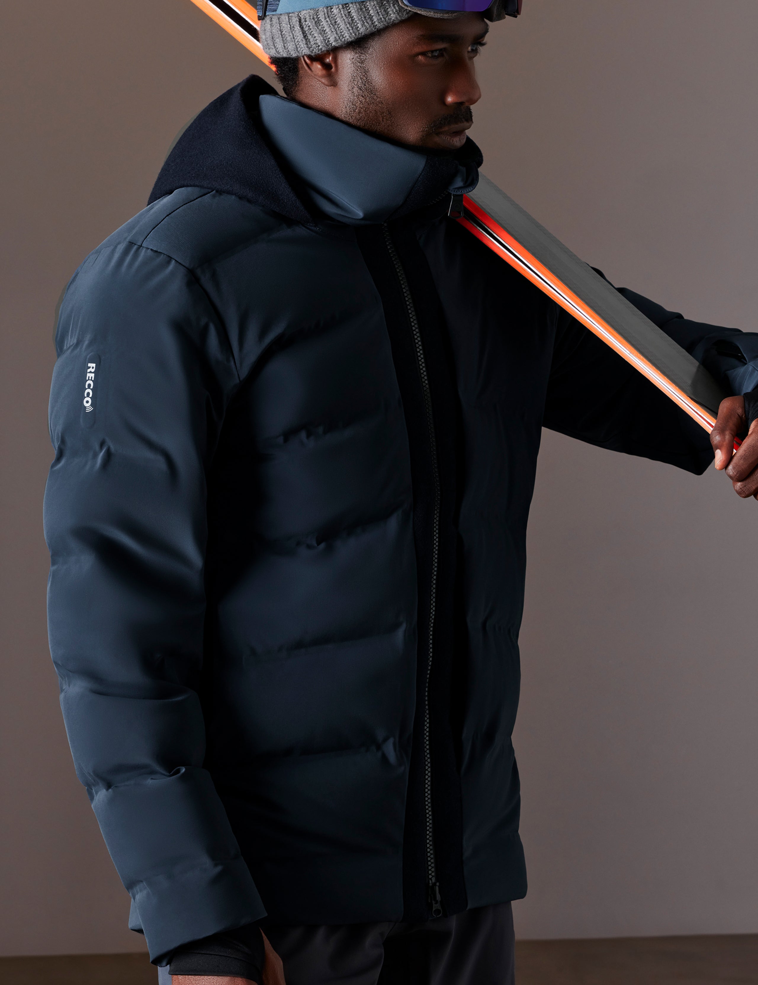 Front 3/4 body view of man holding skis over shoulder wearing Nordic Jacket in Total Eclipse dark blue from AETHER Apparel.