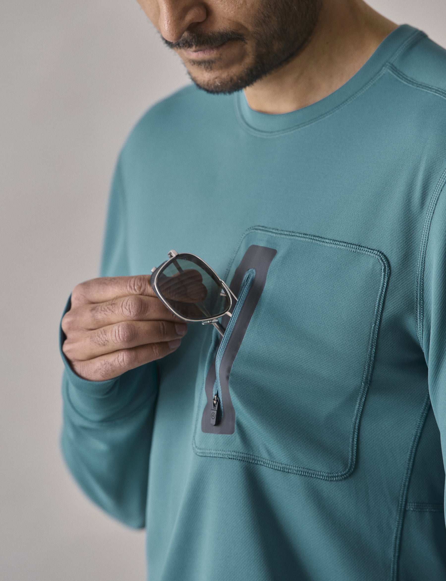 Closeup detailed view of man removing AETHER Summit Sunglass from the zipper-secured offset left chest patch pocket on the Pacific Crewneck Sun Shirt in Aqua Blue from AETHER Apparel.
